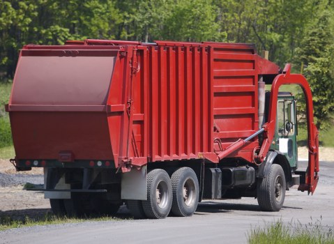 A large red commercial waste collection truck parked on a paved road with a backdrop of green trees and clear sky. The truck features an over-sized rear compaction body with vertical metal ridges, a smooth front cabin with black tires, and a hydraulic loading mechanism on the side. The vehicle is positioned with its rear slightly to the left, and the compacted rubbish container is empty, ready for waste collection. The truck is used for rubbish removal services in Watford, and the image showcases a professional waste management vehicle typical of Commercial Waste Watford’s operations, situated in a semi-rural environment near residential or commercial premises.