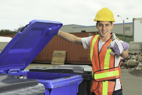 A young male waste management worker wearing a yellow safety helmet, a high-visibility orange and yellow vest, and purple gloves, is standing outdoors near a row of large, open-top blue and black rubbish bins. He is smiling and pointing towards the camera with his right hand while holding the lid of one of the blue bins with his left hand. The background features an industrial or commercial area with warehouses, a stack of wooden pallets, and a partially cloudy sky, indicating a typical rubbish collection or waste removal site. The scene is well-lit with natural daylight, emphasizing the clean, functional appearance of the waste containers and the worker's professional attire. This image aligns with commercial waste removal services offered by companies such as Commercial Waste Watford, serving the Watford area, including postcodes around WD.