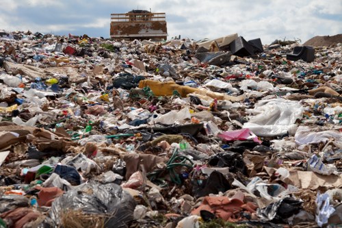 A large, uncontrolled landfill site with a vast accumulation of mixed waste materials, including plastic bags, cardboard, paper, textiles, and other household rubbish scattered across the ground. The waste appears to be in disorganized piles, covering the entire foreground and middle ground of the image, with some items partially buried or overlapped. In the background, there is a partially constructed or abandoned brick building, suggesting an urban or semi-urban environment near Watford, UK. The sky above is partly cloudy, casting diffuse daylight over the scene. This illustration highlights the importance of proper rubbish disposal and waste management services provided by companies like Commercial Waste Watford, especially to prevent environmental pollution and maintain local cleanliness. The overall atmosphere is one of disorder and neglect, emphasizing the need for effective rubbish removal strategies in the area.